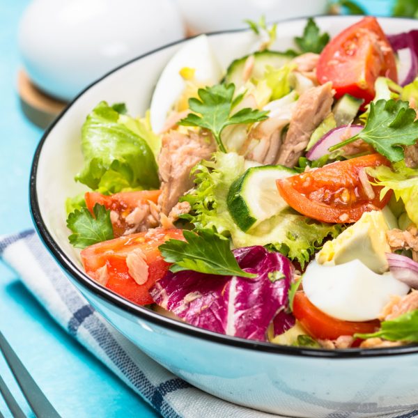 Tuna salad with green leaves, eggs and vegetables in white bowl at blue background.